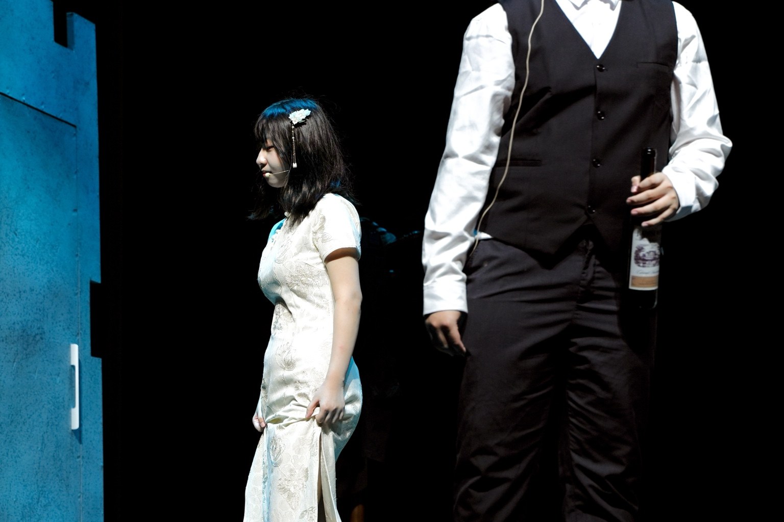 An actor in a light-colored dress beside an actor holding a bottle