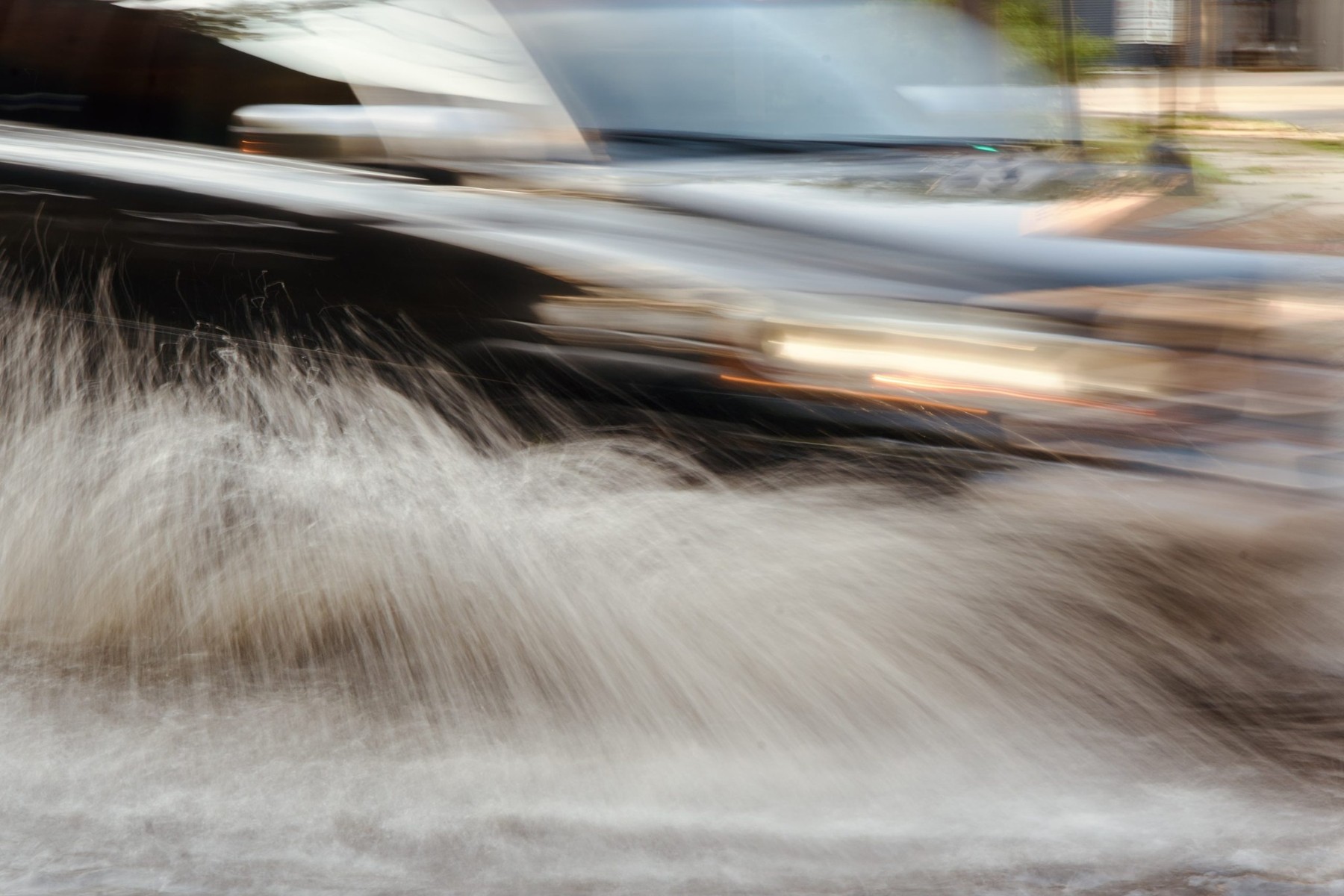 A car drives through a flooded street, spraying water onto the sidewalk.